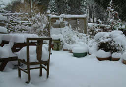 Patio with snow gathering on garden furniture and potted plants.