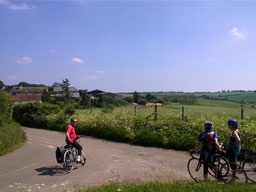 A trio of cyclists stop at a flowery junction looking across a shallow, green valley. A large, wood-framed cottage sits amongst farm buildings to the side.